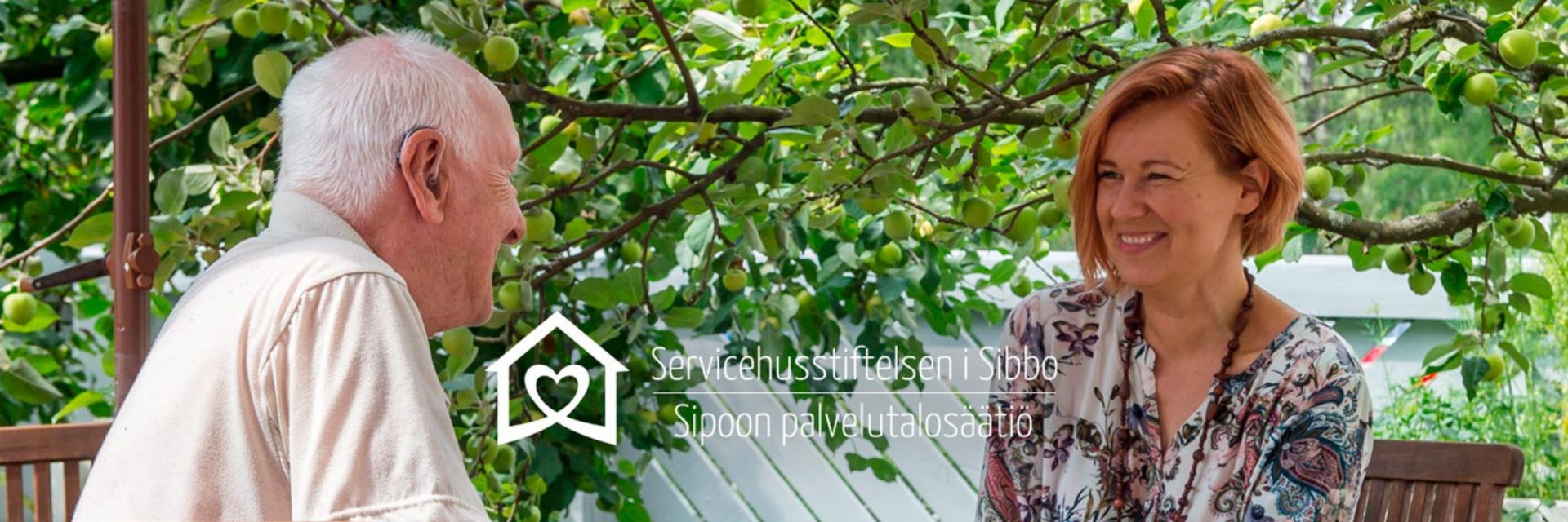 Äldre man och kvinna sitter mitt emot varandra och samtalar under ett träd.